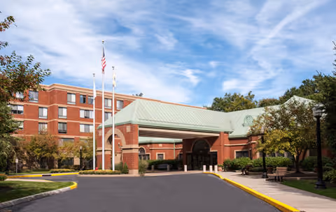 Exterior view of Five Star Premier Residences of Teaneck showing a red brick building with multiple floors, a covered entrance with a green roof, three flagpoles with flags, trees, and a clear blue sky with some clouds.