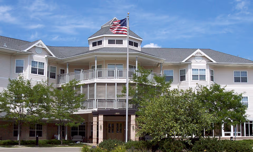 Front facade of a three-story pale siding senior living building with balconies, an American flag on a tall pole, and trees in front.