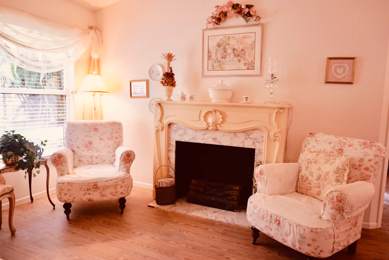 Cozy living room with two floral armchairs flanking an ornate fireplace beneath framed artwork.