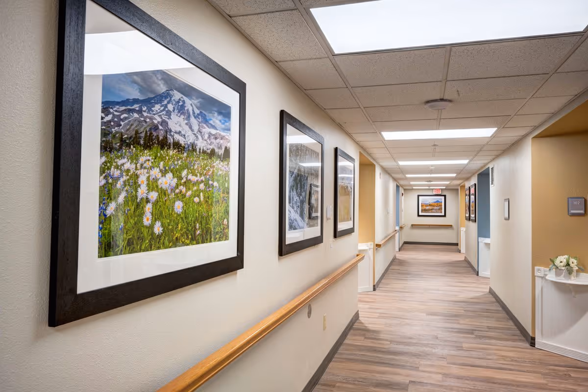 A well-lit hallway in a senior living facility with wooden handrails on both sides and framed landscape photographs hanging on the walls. The floor has wood-like vinyl flooring, and the ceiling has recessed lighting panels. A small table with a flower arrangement is visible on the right side near a door labeled 102.