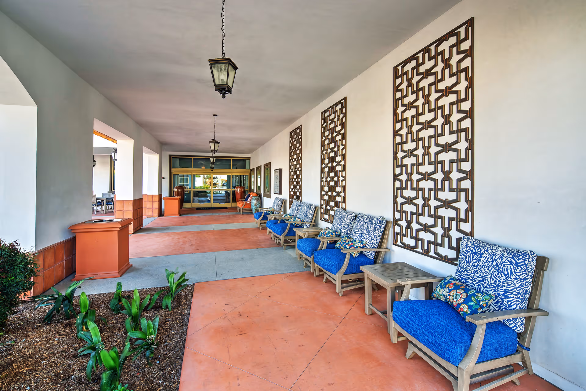 Covered entrance porch with a row of blue-cushioned chairs, decorative wall panels, hanging lanterns, and potted plants.