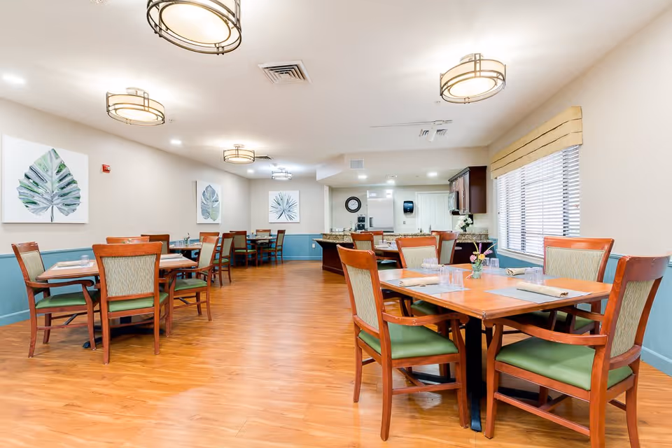A well-lit dining room with wooden floors and several wooden tables surrounded by chairs with green cushions. The walls are painted beige with a blue wainscot, and there are three framed leaf artworks hanging on the walls. The ceiling has modern circular light fixtures, and there is a window with blinds and a beige valance on the right side. In the background, there is a kitchen area with granite countertops and cabinets.