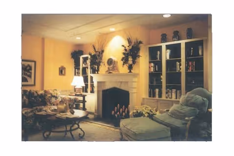 A cozy living room with a fireplace in the center, flanked by built-in bookshelves filled with books and decorative items. There is a floral patterned sofa on the left, a round coffee table with a lamp and decorative items, and a cushioned armchair with an ottoman on the right. The walls are painted a warm yellow color and the room is softly lit with ceiling lights and a table lamp.