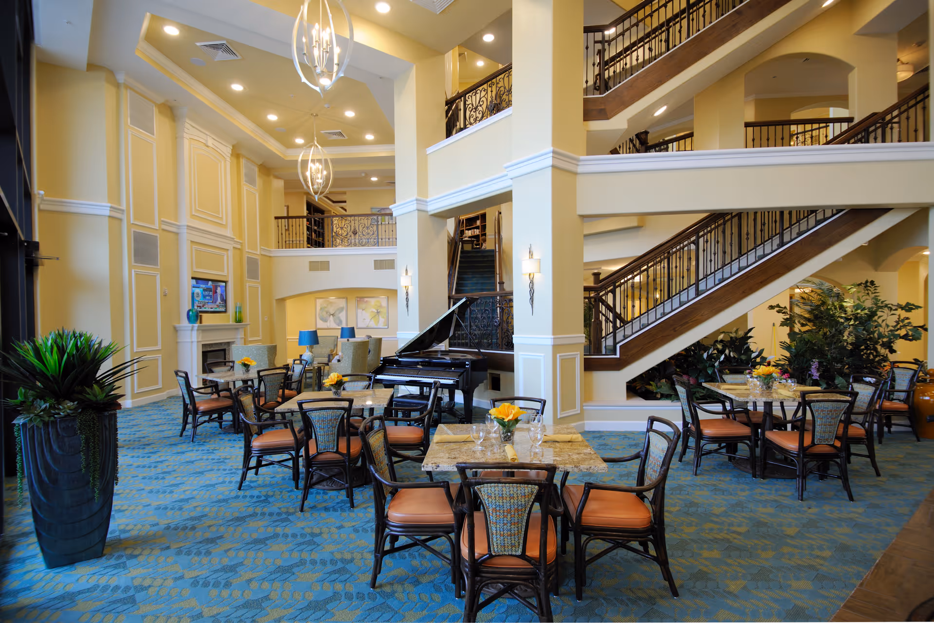 Spacious and elegant senior living facility common area with multiple tables and chairs arranged on a blue patterned carpet. There is a grand piano near the center, a fireplace with a TV above it on the left wall, and a staircase with dark wooden railings leading to an upper floor. The room features high ceilings with hanging light fixtures and large potted plants adding greenery.