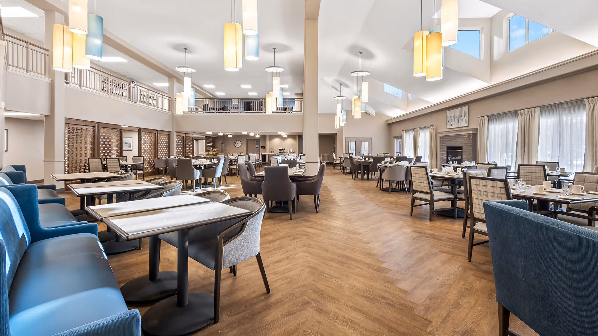 Spacious and well-lit dining room with multiple tables and chairs arranged neatly. The room features high ceilings with hanging pendant lights, large windows with curtains, and a fireplace on one side. The floor has a wood pattern, and there is a second-floor balcony overlooking the dining area.