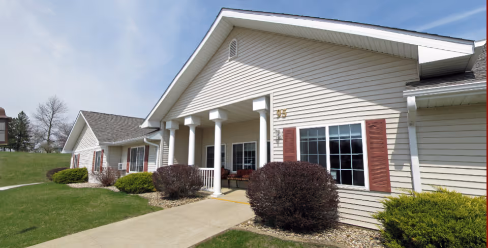 Exterior view of Arlington Place at Grundy Center, showing a single-story building with beige siding, white columns supporting a covered entrance, red shutters on windows, and neatly trimmed bushes along a concrete walkway.