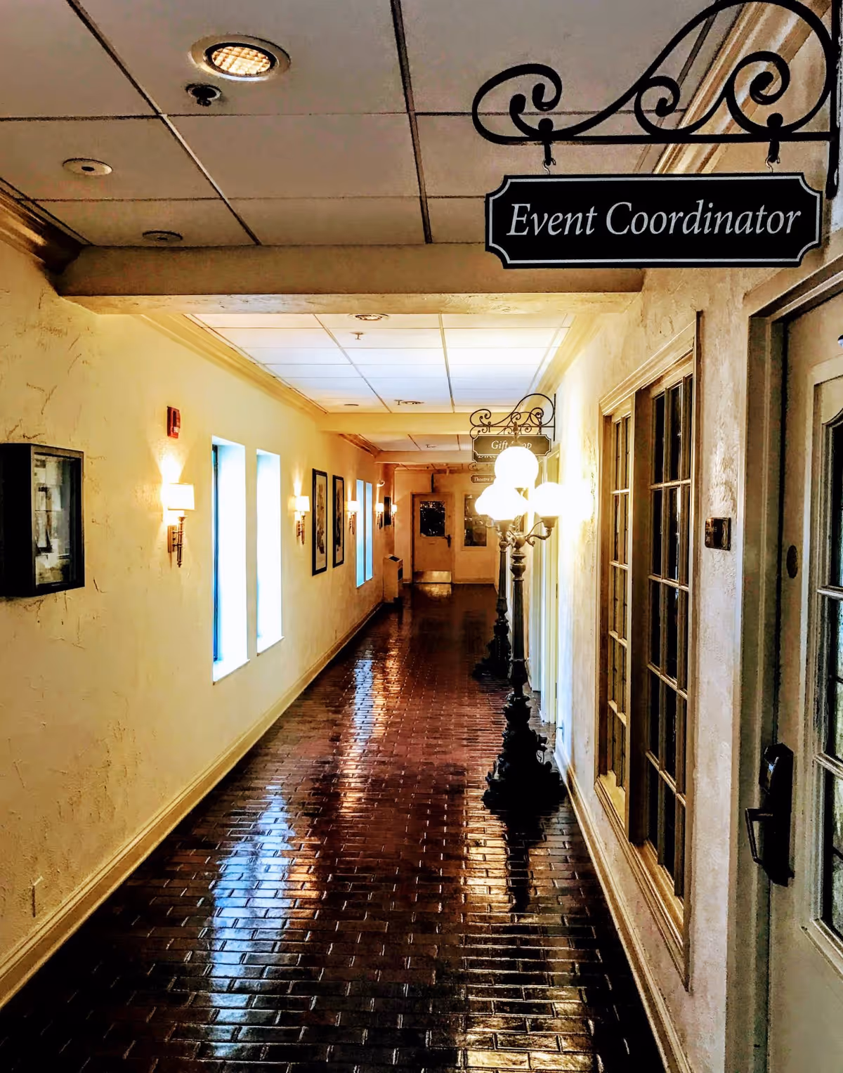 A long, narrow hallway with shiny dark tile flooring and cream-colored textured walls. The ceiling has recessed lighting and a drop ceiling. On the right side, there are vintage-style street lamp posts with glowing lights and a door with glass panels. Hanging from the ceiling is a decorative black sign that reads 'Event Coordinator.' The left wall has small windows and framed pictures with wall-mounted sconces providing additional light.