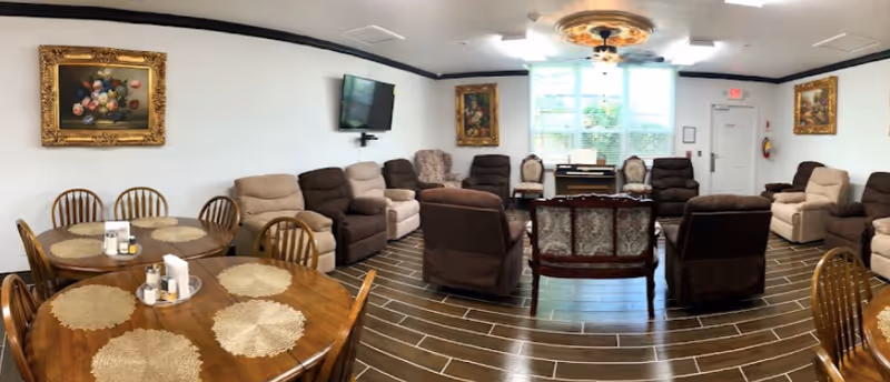 A spacious living room in an assisted living facility featuring multiple comfortable recliner chairs arranged in a semi-circle facing a wall-mounted flat screen TV. There are two round wooden tables with chairs and placemats in the foreground. The room has large windows with blinds, framed floral paintings on the walls, and a decorative ceiling light fixture. The floor is covered with wood-look tiles.