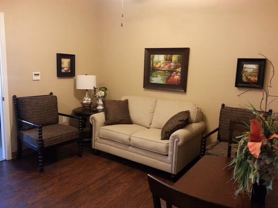 A cozy living room with a beige sofa and two dark wooden chairs with brown cushions. There is a small round table with a lamp and decorative items between the sofa and one chair. Three framed paintings hang on the beige walls. A floral arrangement is partially visible on the right side of the image.
