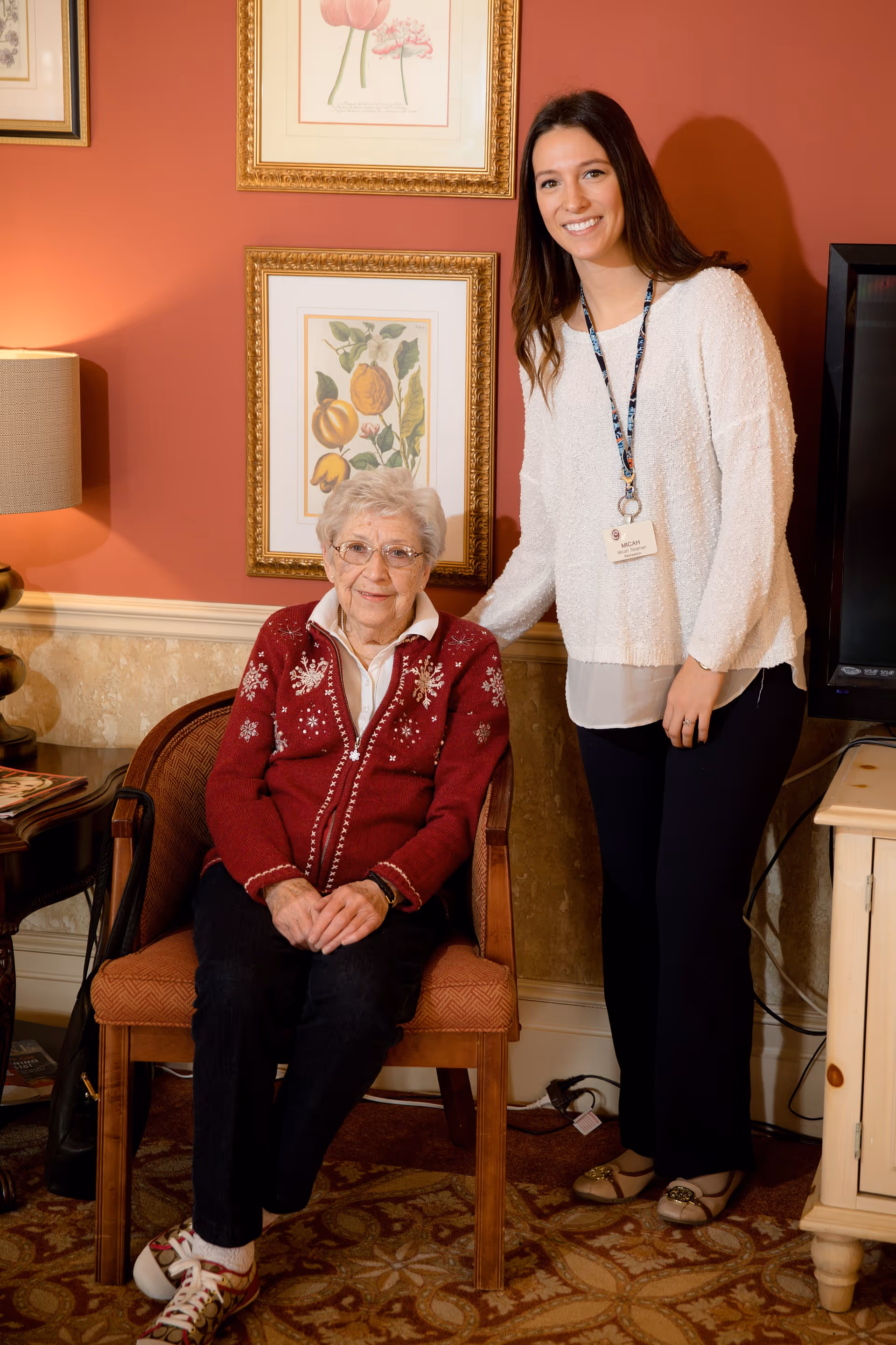 An elderly woman wearing a red sweater with white patterns sits on a wooden chair in a warmly decorated room. A younger woman with long brown hair, dressed in a white sweater and black pants, stands beside her smiling. The room has framed botanical prints on the wall, a table lamp, and a patterned carpet.