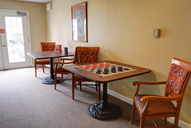A small seating area with two wooden tables and four chairs with patterned cushions. One table has a checkers board with red and black pieces set up. The room has beige walls, a carpeted floor, a framed picture on the wall, and a door with glass panels leading outside.