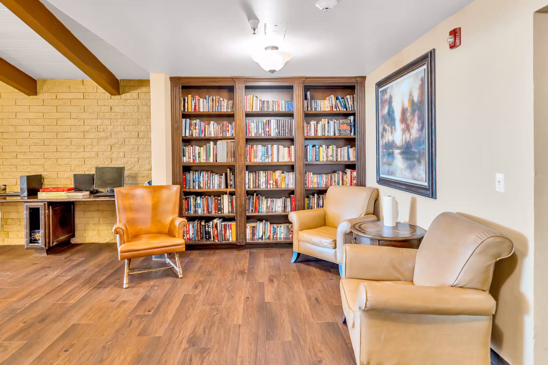 A cozy reading area in a senior living facility with three leather armchairs arranged around a small round wooden table. Behind the chairs is a large wooden bookshelf filled with books. To the left, there is a desk with two computers and some board games. The room has wooden flooring, a beige wall with a framed painting, and a yellow brick accent wall with exposed wooden beams on the ceiling.