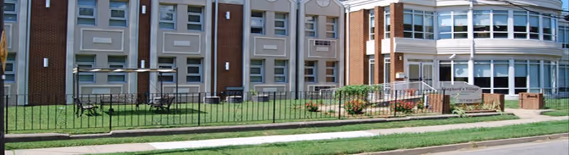 Exterior view of Shepherd's Village At Park Avenue, showing a multi-story brick and white building with large windows, a fenced grassy area with benches and tables, and a sign with the facility's name near the entrance.