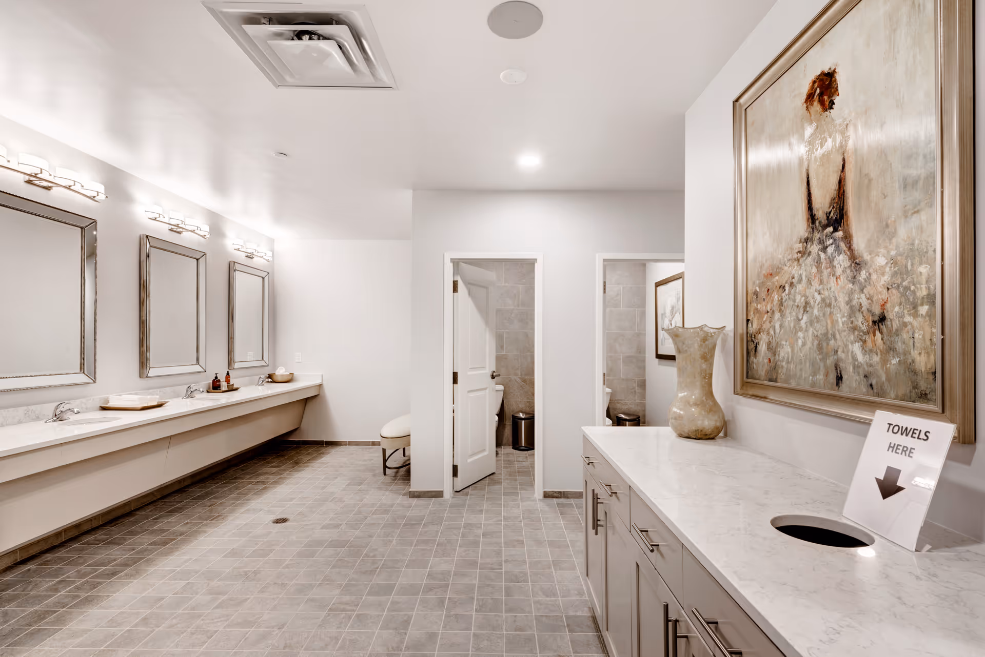 A spacious, modern bathroom with three large mirrors and sinks on the left wall, two open toilet stalls in the center, and a countertop with a vase and a sign indicating towel disposal on the right. The floor is tiled in gray, and the walls are painted white. A large painting of a woman in a dress hangs above the countertop.