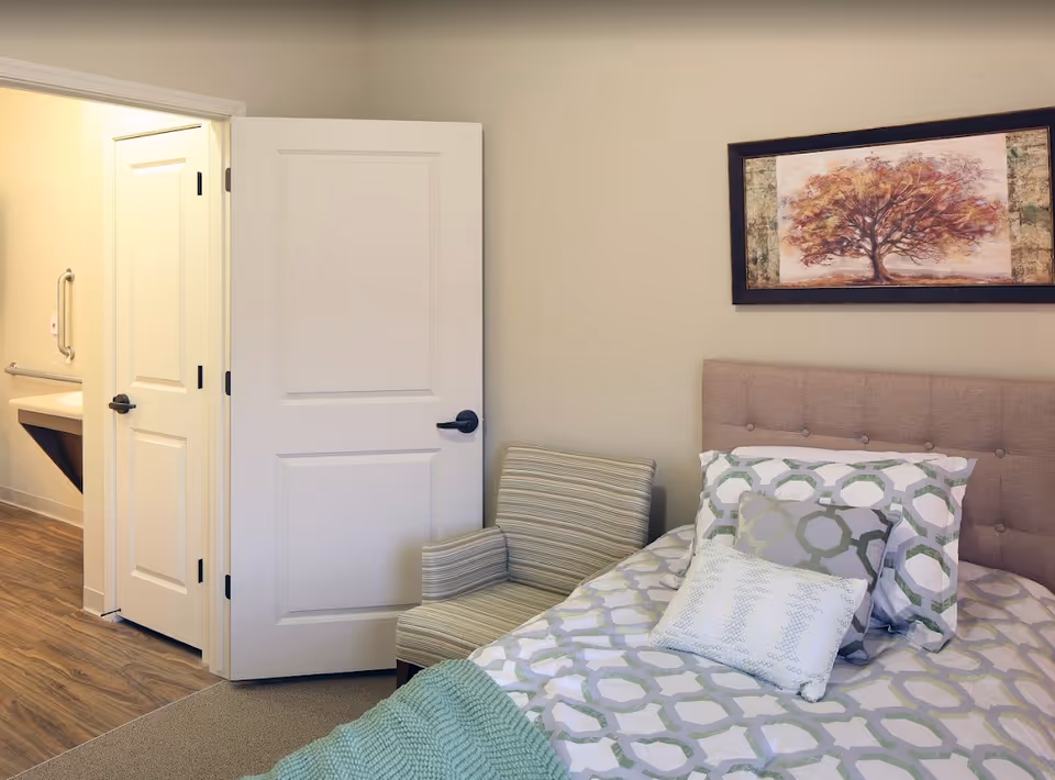 A bedroom in a senior living facility featuring a bed with patterned bedding and multiple pillows, a striped armchair next to the bed, a framed painting of a tree on the wall, and an open door leading to a bathroom with a sink and grab bars.