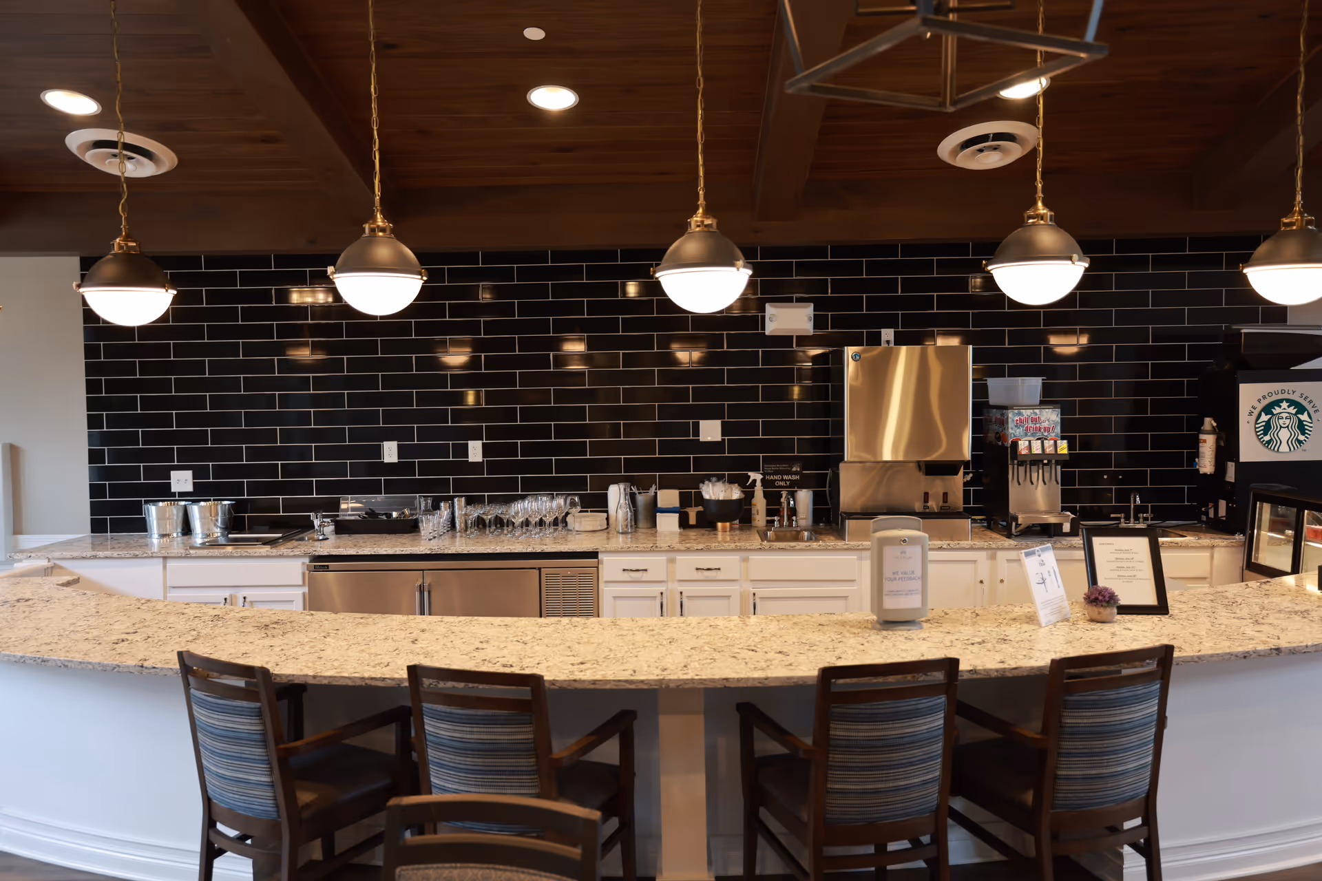 A modern kitchen or cafe counter area with a curved granite countertop and four wooden chairs with striped cushions. Above the counter, there are five hanging pendant lights with round white shades. The back wall is covered with glossy black subway tiles and features various kitchen appliances, glassware, and a Starbucks coffee machine.