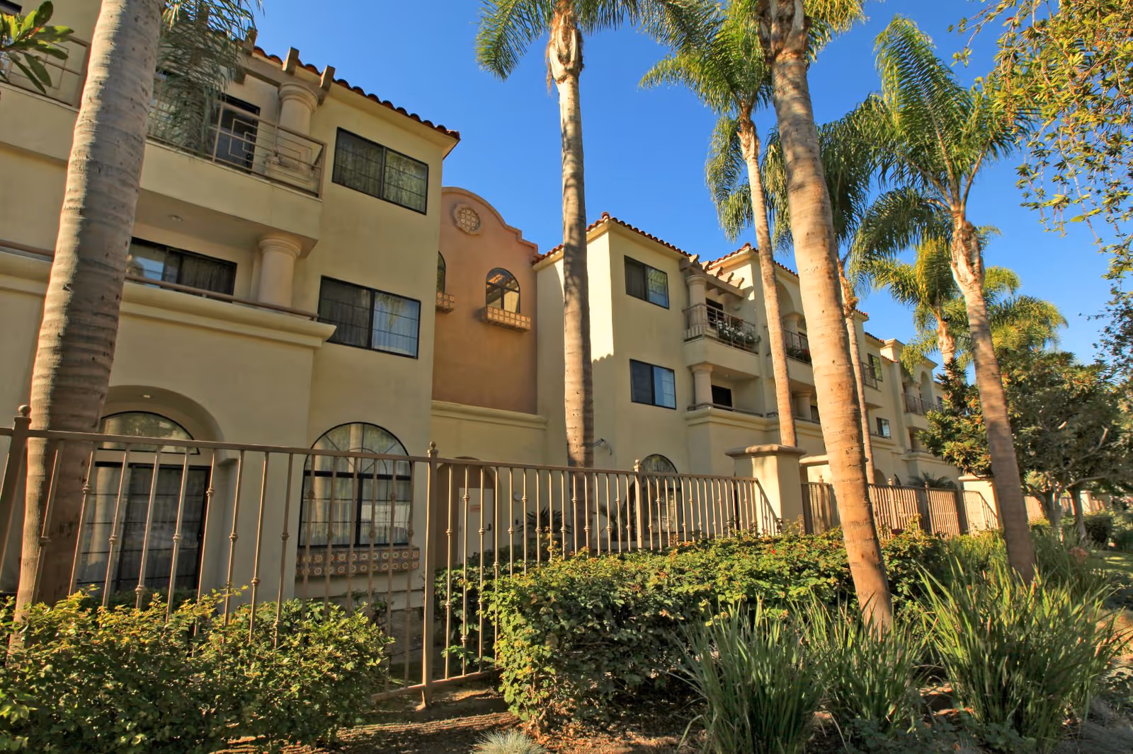 Exterior view of a multi-story residential building with beige stucco walls and red tile roof, surrounded by palm trees and greenery under a clear blue sky.