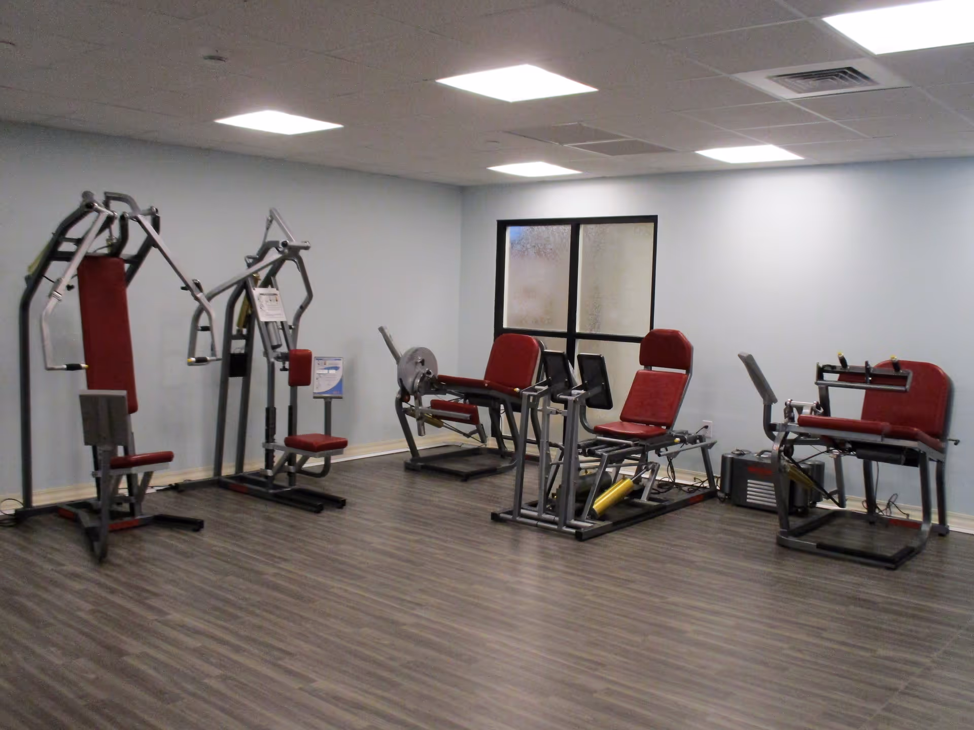 An interior fitness room with several red-padded exercise machines against a pale blue wall.