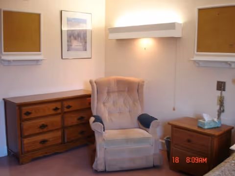 A cozy corner of a room featuring a beige upholstered armchair with dark armrest covers, positioned between a wooden dresser with six drawers on the left and a wooden nightstand with a tissue box on top on the right. The walls are light-colored with two empty cork bulletin boards and a framed picture hanging above the dresser. A wall-mounted light fixture is above the armchair.
