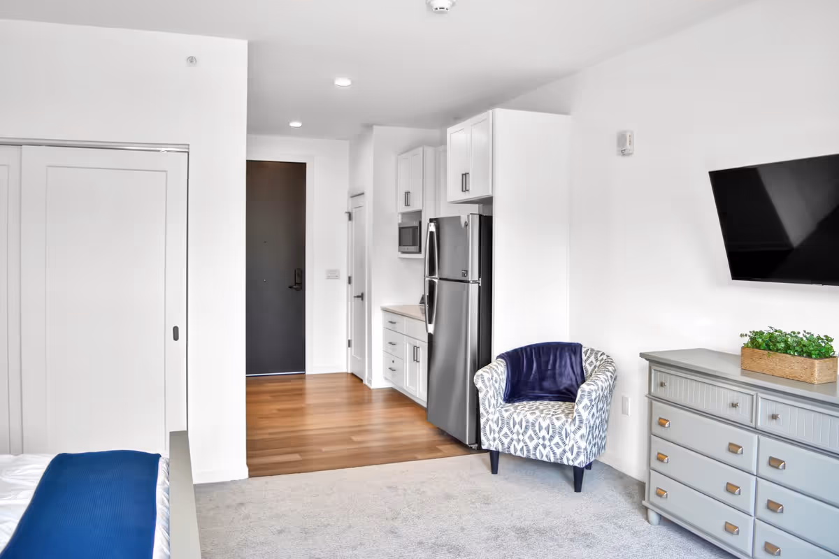 A modern studio apartment interior featuring a small kitchen area with white cabinets, a stainless steel refrigerator, and a microwave. There is a patterned armchair with a dark blue throw blanket, a gray dresser with a green plant on top, and a wall-mounted flat-screen TV. The floor transitions from carpet in the living area to wood in the kitchen and entryway. A closed white sliding door and a dark entry door are visible in the background.