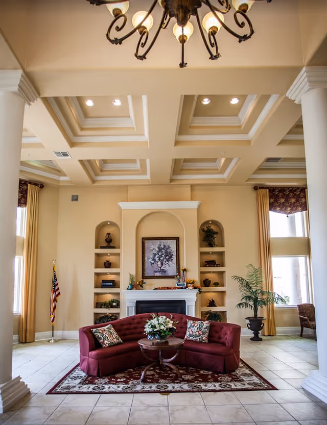 A spacious and elegant living room area with a burgundy curved sofa adorned with floral pillows, centered on a patterned rug. A round wooden coffee table with a floral arrangement sits in front of the sofa. The room features a decorative fireplace with built-in shelves on either side holding various decorative items and plants. Large windows with gold curtains allow natural light to fill the space. The ceiling has a coffered design with recessed lighting and a chandelier hanging above. An American flag stands in the corner near one window.