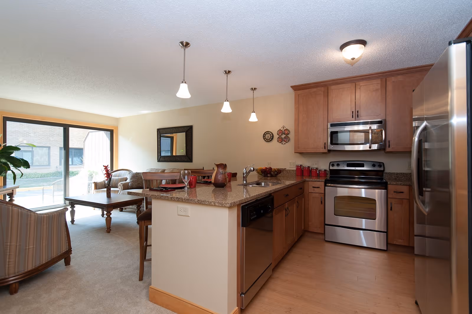 A modern kitchen and living room area in a senior living community. The kitchen features wooden cabinets, stainless steel appliances including a refrigerator, oven, microwave, and dishwasher, and a granite countertop island with a sink and bar stools. The living room has a coffee table, armchairs, a sofa, and a large window with sliding glass doors letting in natural light.