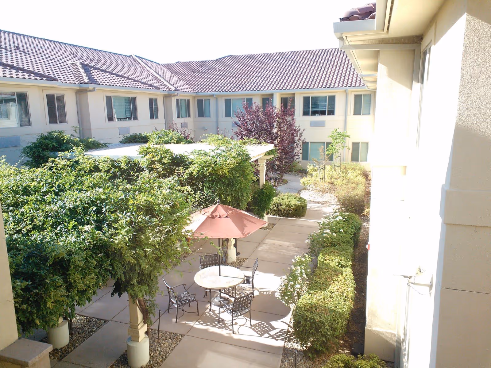 Outdoor courtyard area at Westmont of Brentwood featuring a round table with four metal chairs and a closed red umbrella. The courtyard is surrounded by green bushes, trees, and a two-story building with beige walls and multiple windows.