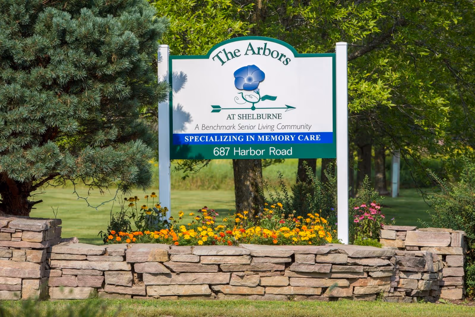 Outdoor sign for The Arbors at Shelburne, a senior living community specializing in memory care, surrounded by flowers and greenery with trees in the background.