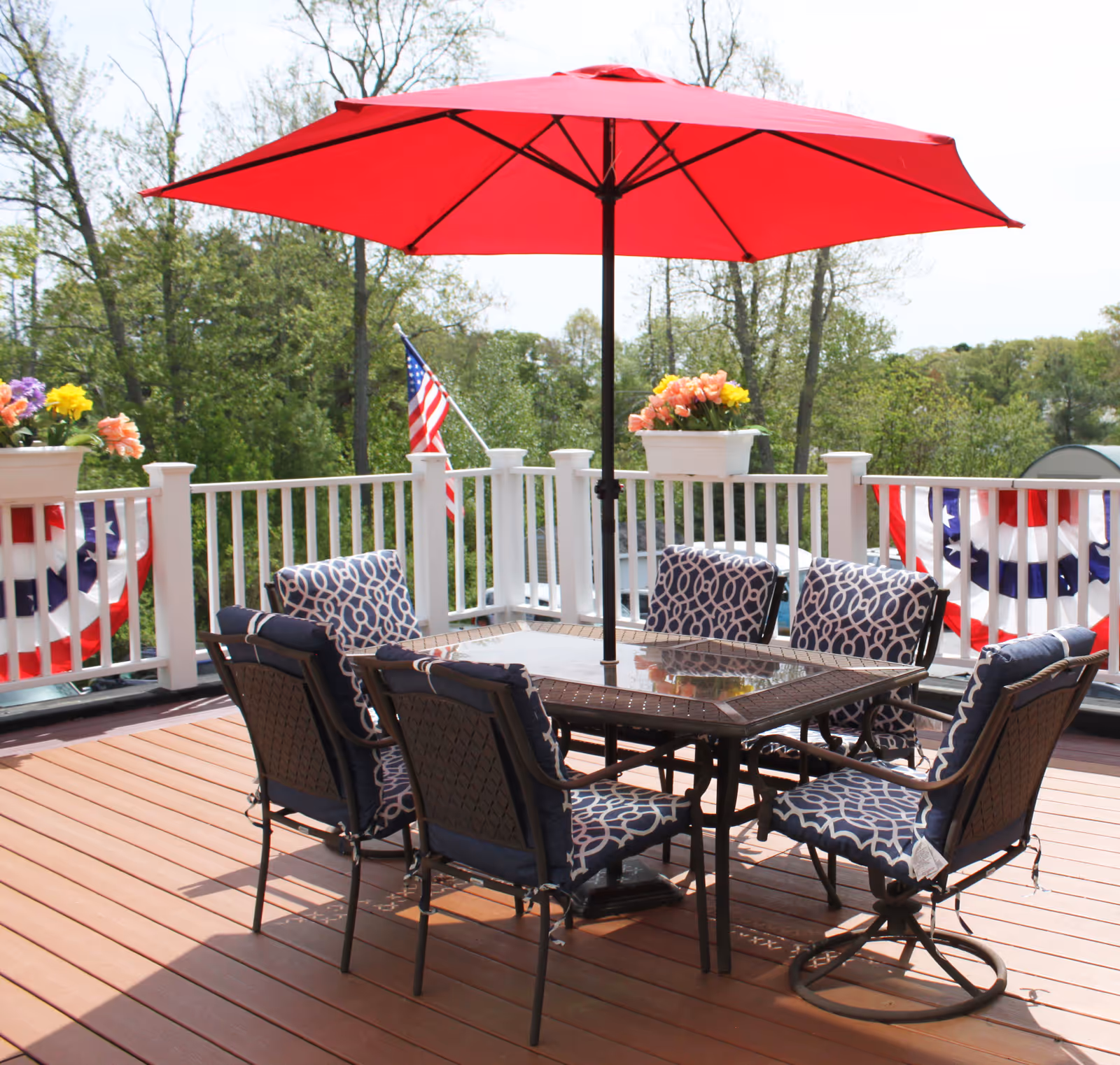 Sunlit outdoor deck with a dining table surrounded by six patterned chairs under a red umbrella, with flower planters and an American flag on the railing.