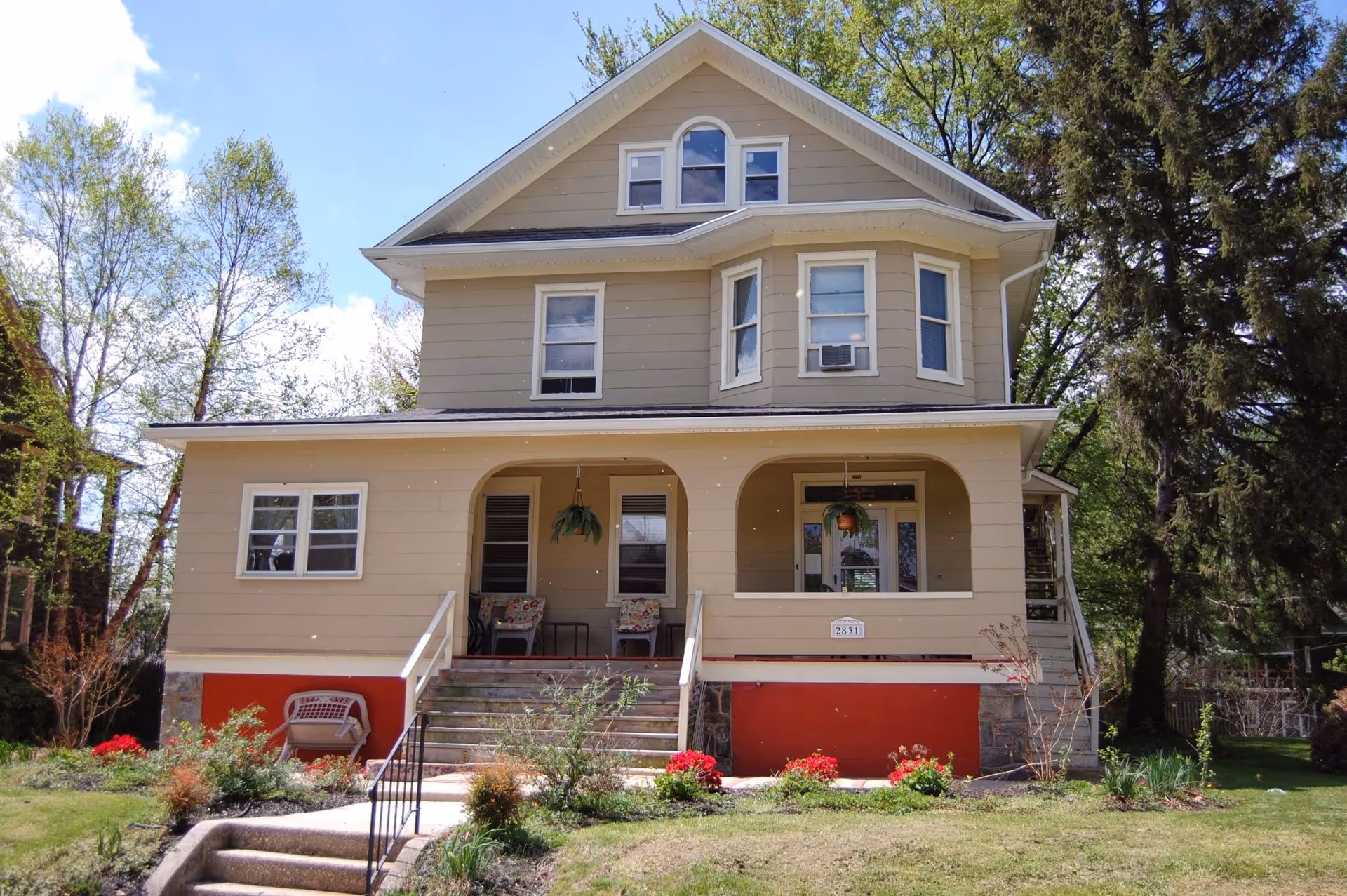 Three-story beige house with a covered front porch, steps up from a lawn and trees surrounding the property.