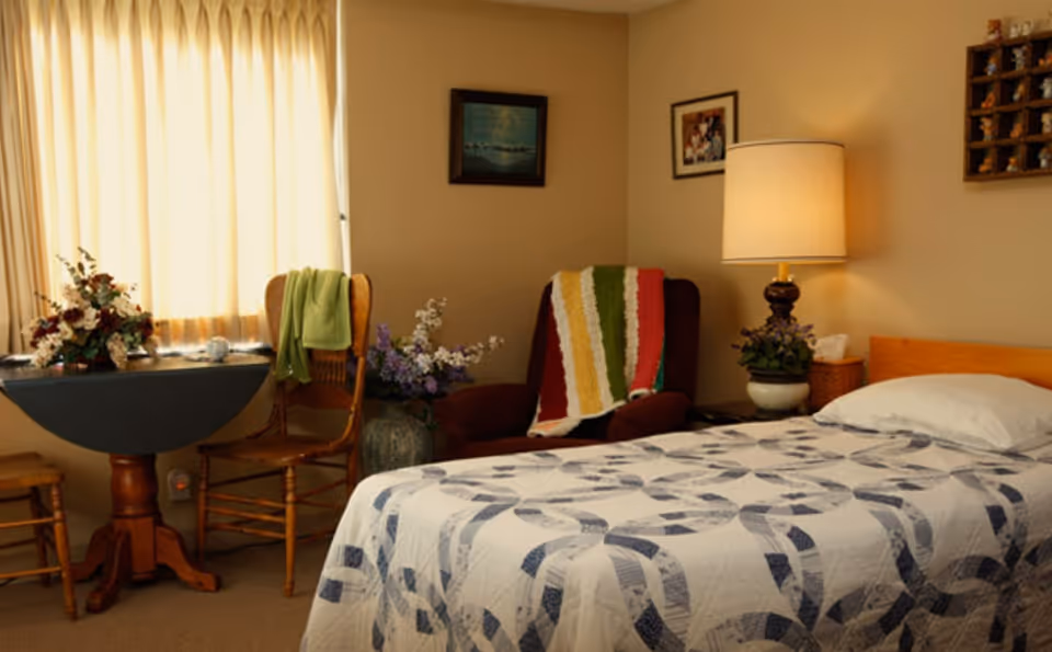 Single-occupancy bedroom with a patterned quilted bed, an armchair draped with a striped blanket, a side table and lamp, and a curtained window.