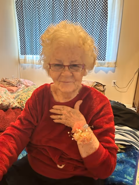 An elderly woman with curly white hair and glasses wearing a red sweater sits on a bed in a room. She has a floral corsage on her wrist and is touching her chest with one hand. Behind her, there are patterned curtains covering a window and various blankets and clothes on the bed.