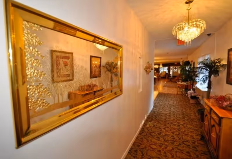 Carpeted interior hallway in an assisted living facility with a large gold-framed mirror, chandeliers, plants, and decorative furniture.