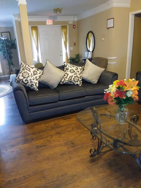 A cozy living room area with a dark gray sofa adorned with four patterned throw pillows. In front of the sofa is a glass coffee table with a metal frame, holding a vase filled with colorful flowers. The room has wooden flooring, beige walls, and a white door with yellow curtains in the background. There is a round mirror on the wall and a chair next to it.