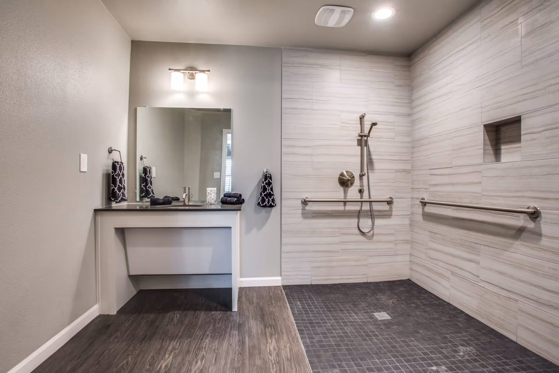 A modern bathroom featuring a walk-in shower with a handheld showerhead and two horizontal grab bars mounted on tiled walls. The floor of the shower area is covered with dark tiles, while the rest of the bathroom has wood-like flooring. A vanity with a black countertop, a sink, a large mirror, and a light fixture above is visible on the left side. Black and white patterned hand towels are hanging on towel rings near the sink and shower.