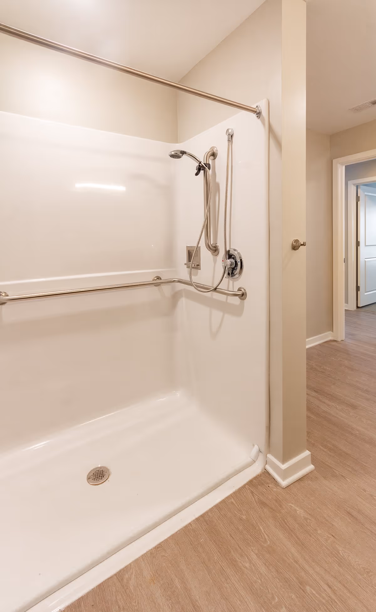 A clean, accessible shower area with grab bars and a handheld showerhead in a senior living facility. The shower has a white surround and a metal curtain rod above. The floor outside the shower is wood-patterned, and a hallway with open doors is visible in the background.