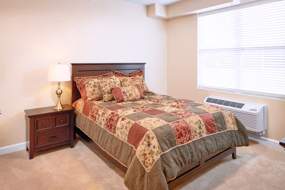 Well-lit bedroom with a patterned quilted bed, wooden headboard and nightstand with lamp beside a large window.