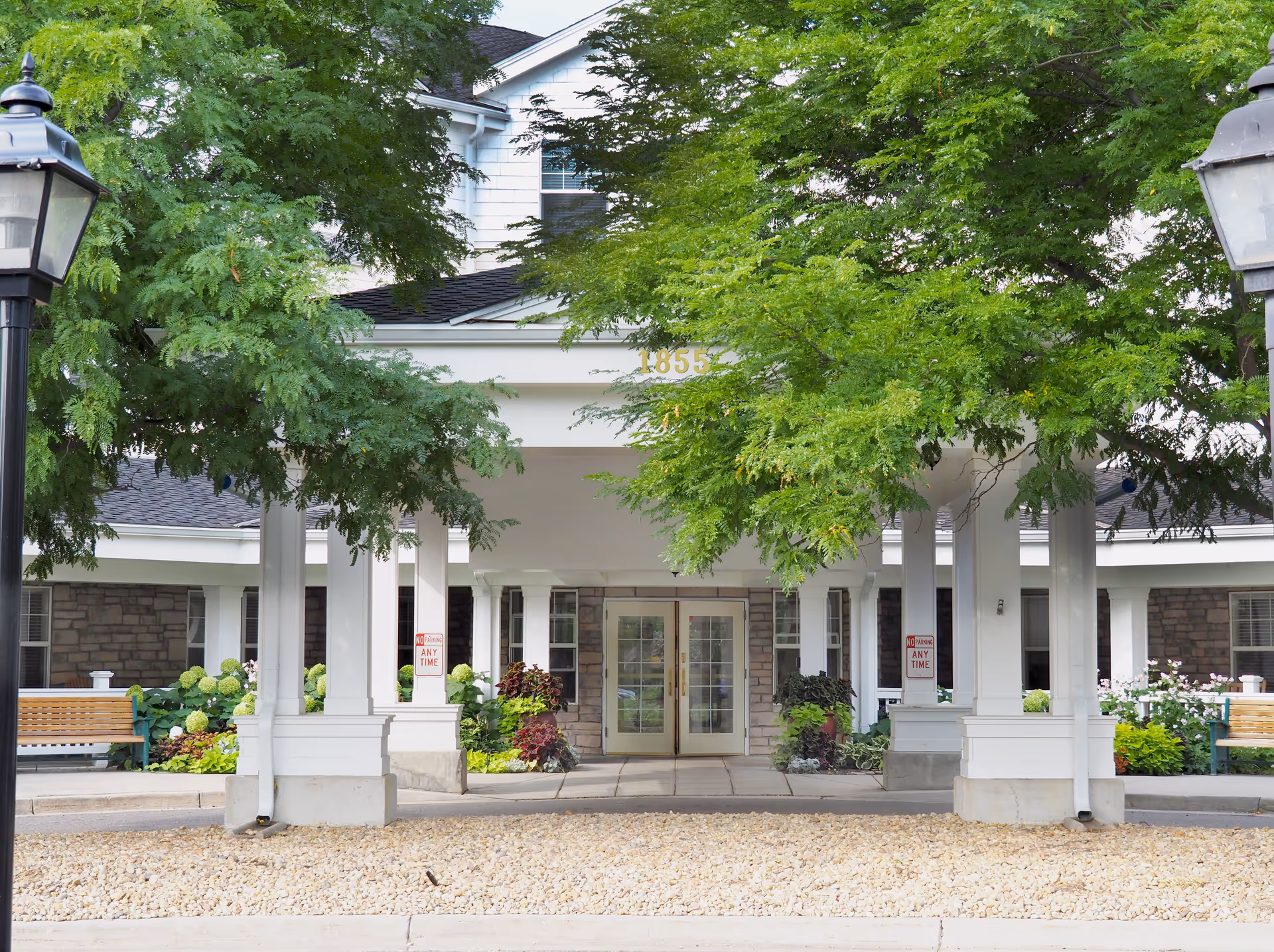 Entrance of Balfour Retirement Community featuring a covered driveway with white pillars, green leafy trees partially obscuring the building number 1855, stone walls, glass double doors, benches, and landscaped plants.