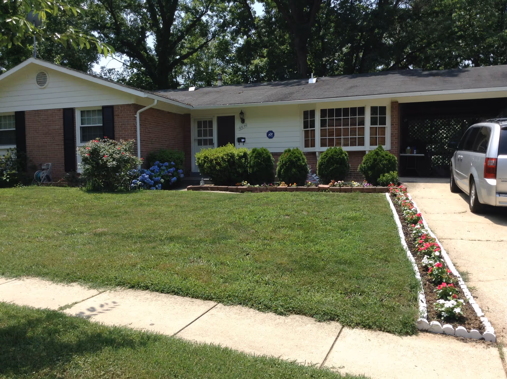 Front exterior view of a single-story residential care home with a well-maintained lawn, flower beds along the driveway, bushes near the entrance, and a silver minivan parked in the driveway.