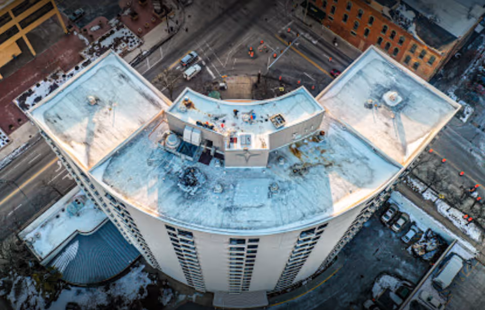 Aerial view of a curved multi-story building's snow-dusted rooftop and upper facade surrounded by city streets and parked cars.