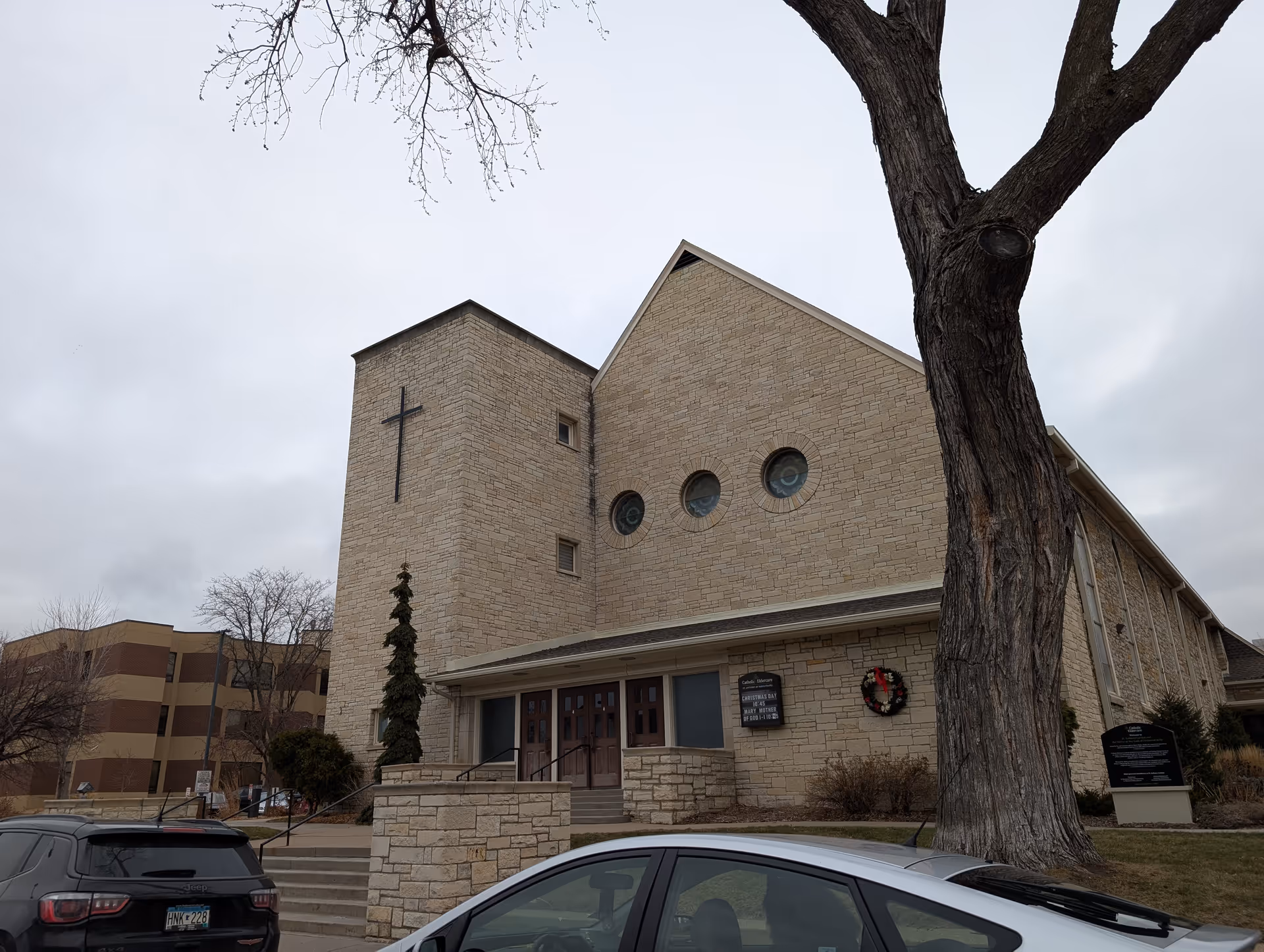 Exterior view of a stone building with a cross mounted on a tower section, three circular stained glass windows, and a wreath on the wall near the entrance. There are cars parked in front and a large tree on the right side. The sky is overcast.
