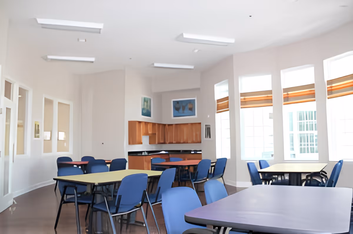 A bright and spacious dining room with multiple tables and blue chairs arranged around them. The room features large windows with brown blinds, light-colored walls, and a small kitchenette area with wooden cabinets and a black countertop. The ceiling has several rectangular fluorescent lights.