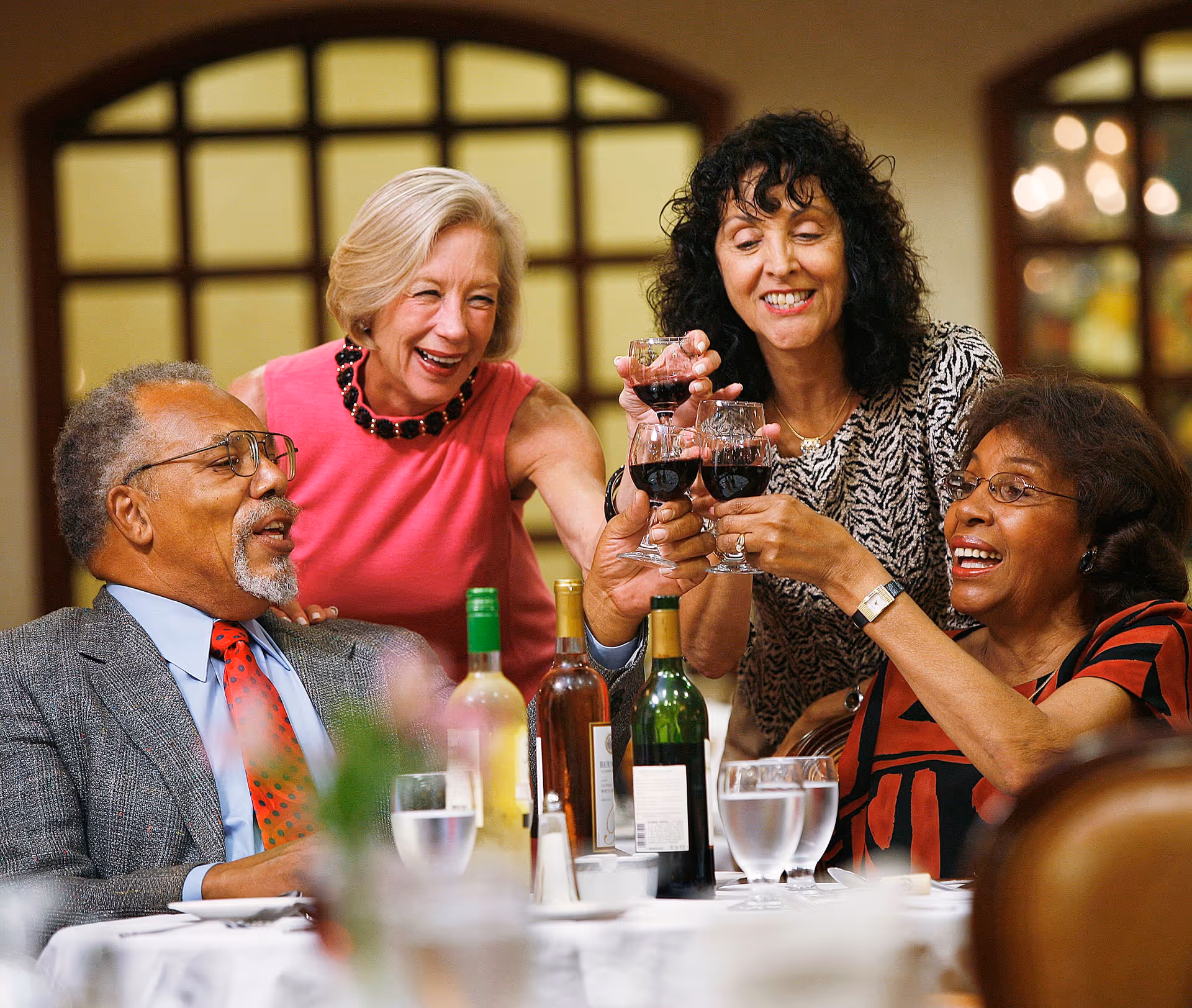 Four older adults sitting around a dining table, smiling and raising glasses of red wine in a toast. The table has bottles of wine and glasses of water, and the background shows a warmly lit interior with arched windows.