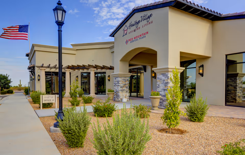 Front entrance of a beige assisted living building with arched entry, landscaped gravel beds, planters, a flagpole, and a walkway.