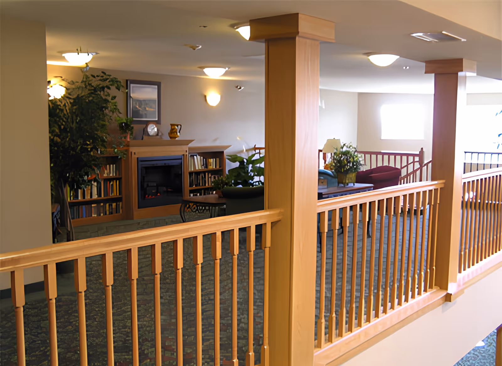 Interior view of a senior living facility lounge area with wooden railings, columns, a fireplace surrounded by bookshelves, potted plants, and seating including chairs and a table with a lamp and flower arrangement.