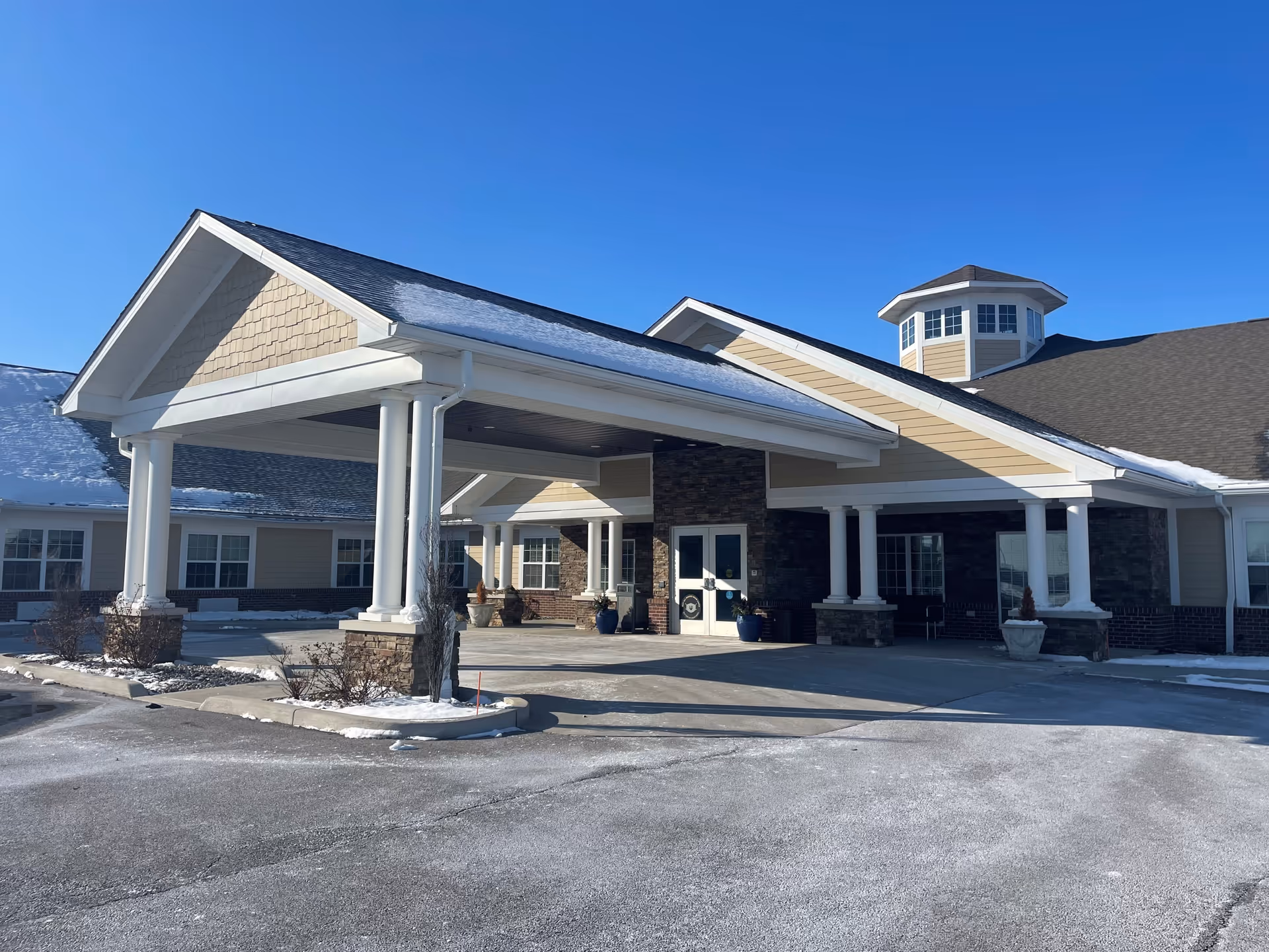 Exterior view of The Lakes of Sylvania senior living facility entrance with a covered drop-off area, columns, and a clear blue sky. There is some snow on the ground and roof.