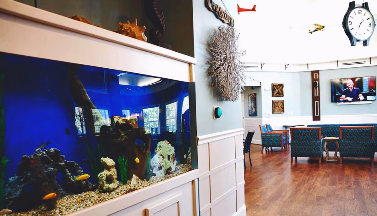 A bright senior living common area showing a large built-in aquarium on the left, seating chairs and tables, and a wall-mounted TV.