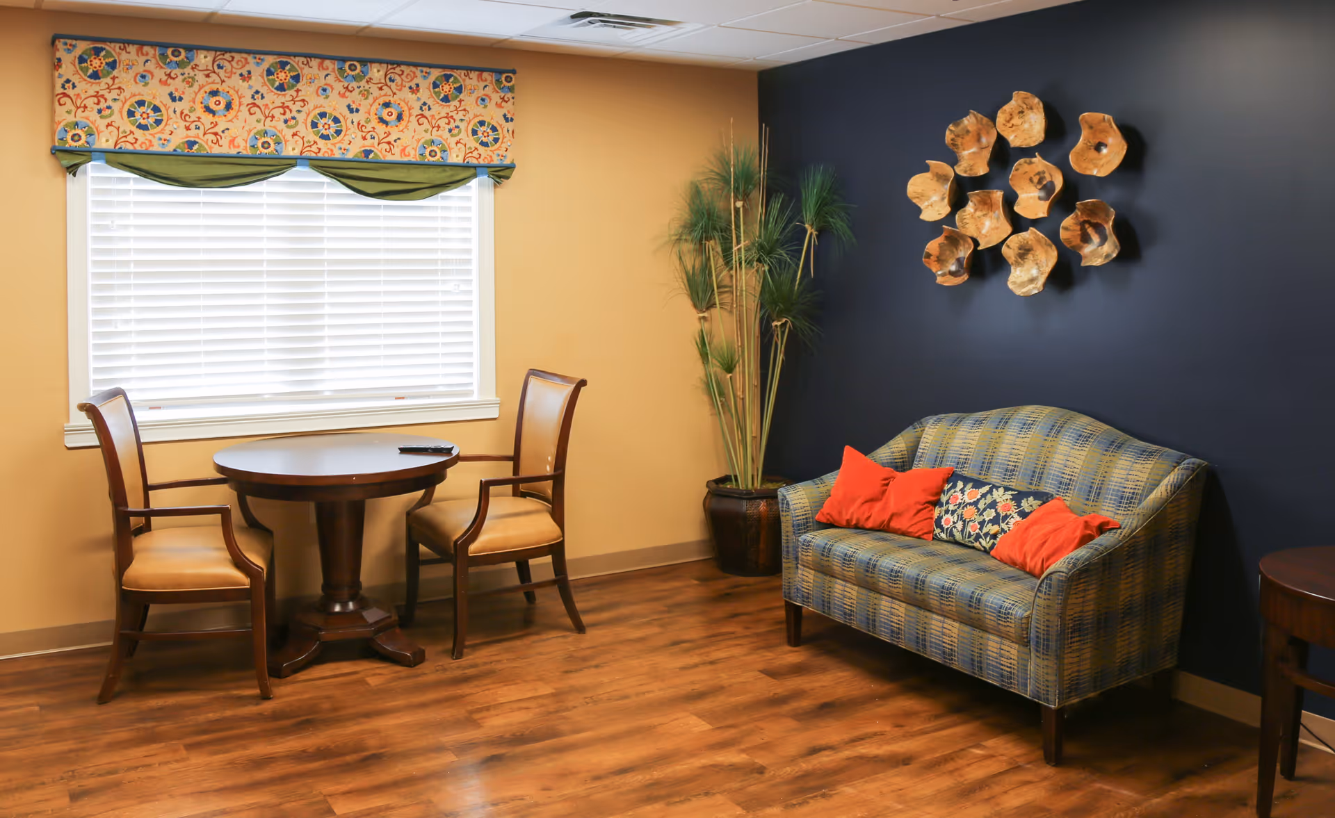 A cozy sitting area with a small round wooden table and two wooden chairs with beige cushions near a window with white blinds and a colorful valance. To the right, there is a blue patterned loveseat with three decorative pillows, two red and one floral. A tall potted plant is placed in the corner against a dark blue wall adorned with wooden wall art resembling abstract flowers.