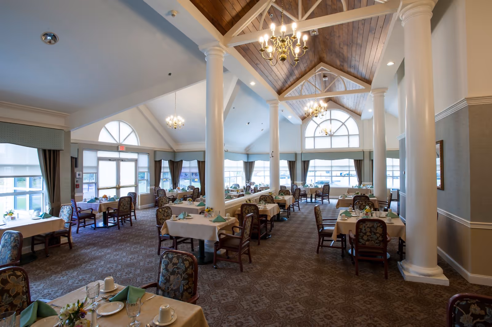 A spacious dining room with multiple tables covered in beige tablecloths, each set with green napkins, plates, cups, and small flower arrangements. The room features large arched windows, tall white columns, chandeliers hanging from a vaulted wooden ceiling, and patterned carpet flooring.