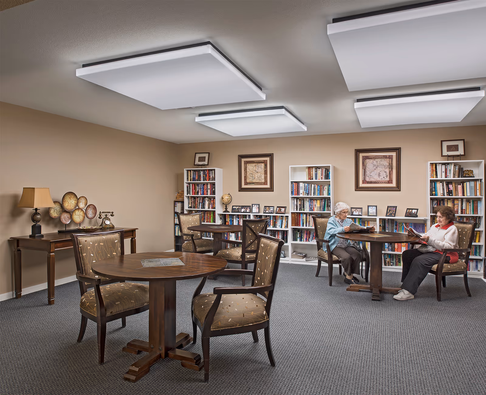 A cozy reading room in a senior living facility with two elderly women sitting at a round wooden table reading magazines. The room has beige walls, carpeted floor, several bookshelves filled with books, framed pictures on the walls, and decorative items on a side table including a lamp and a vintage telephone.